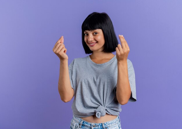 Smiling young brunette caucasian girl gestures money hand sign with two hands isolated on purple background with copy space