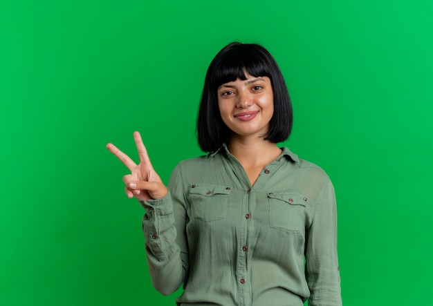 Smiling young brunette caucasian girl gestures victory hand sign isolated on green background with copy space