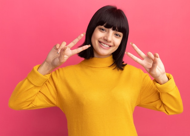 Smiling young brunette caucasian girl gestures victory sign with hands on pink