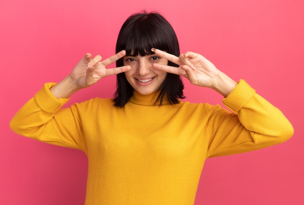 Smiling young brunette caucasian girl gesturing victory sign looking at camera through fingers isolated on pink wall with copy space