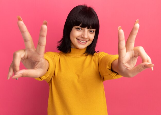 Smiling young brunette caucasian girl gesturing victory sign on pink