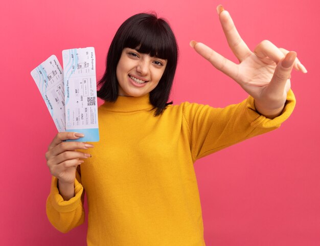 Smiling young brunette caucasian girl holds air tickets and gestures victory hand sign