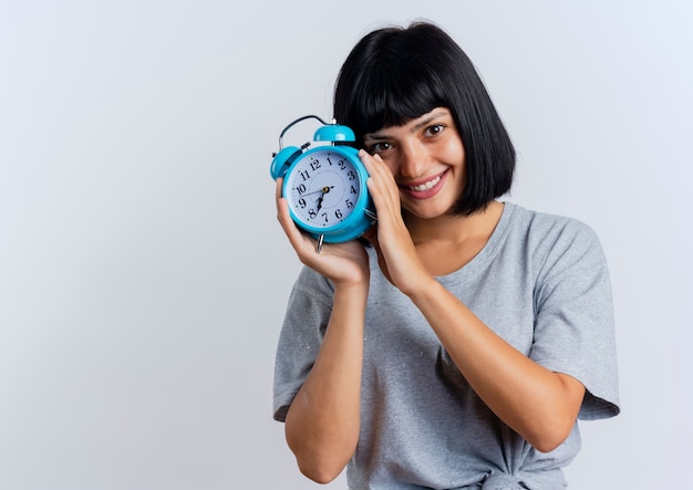 Smiling young brunette caucasian girl holds alarm clock looking  