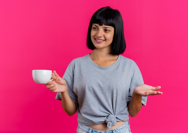 Foto gratuita la giovane ragazza caucasica castana sorridente tiene la tazza e guarda al lato isolato su fondo rosa con lo spazio della copia