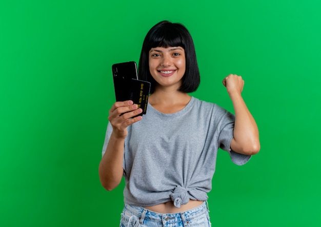 La giovane ragazza caucasica castana sorridente tiene il telefono e la carta di credito che mantengono il pugno isolato su fondo verde con lo spazio della copia