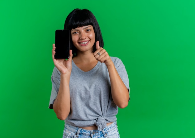 Smiling young brunette caucasian girl holds phone and thumbs up isolated on green background with copy space