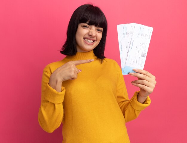 Smiling young brunette caucasian girl holds and points at air tickets
