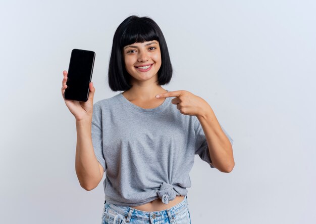 Smiling young brunette caucasian girl holds and points at phone 