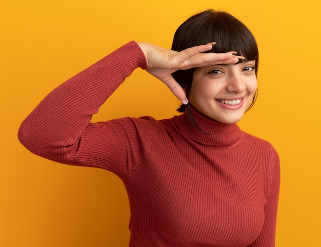 Smiling young brunette caucasian girl keeps palm at forehead isolated on orange wall with copy space