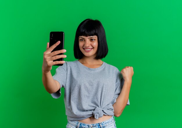 Smiling young brunette caucasian girl looks at phone and keeps fist isolated on green background with copy space