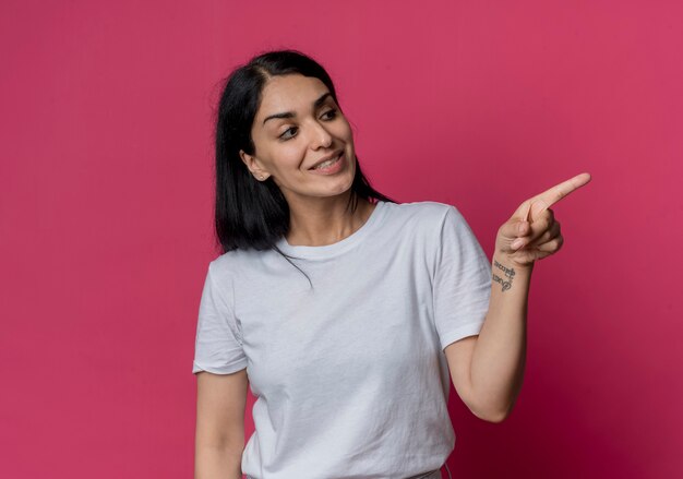 Smiling young brunette caucasian girl looks and points at side isolated on pink wall