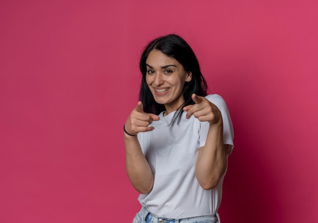 Smiling young brunette caucasian girl points with two hands isolated on pink wall
