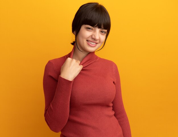 Smiling young brunette caucasian girl pulls down collar and looks at camera on orange