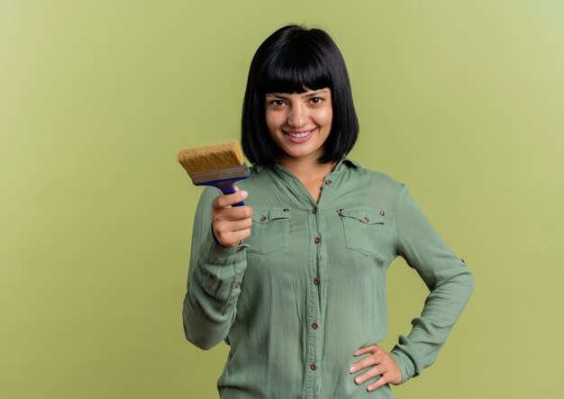 Smiling young brunette caucasian girl puts hand on waist and holds paint brush looking at camera isolated on olive green background with copy space