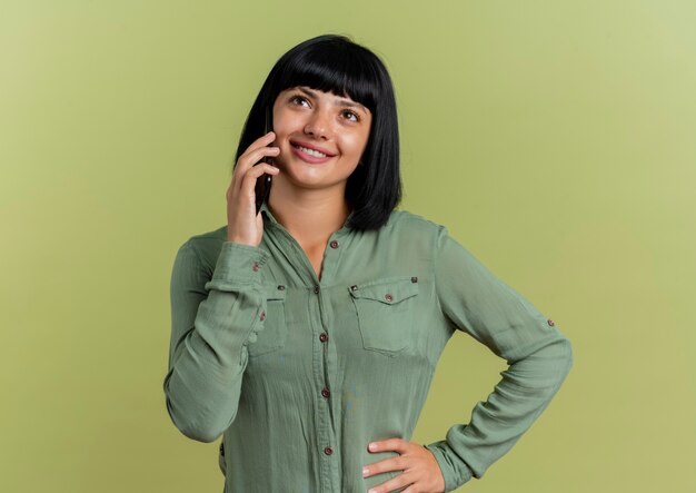 Smiling young brunette caucasian girl puts hand on waist talking on phone looking up 