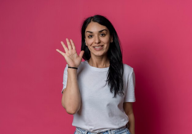 Smiling young brunette caucasian girl raises hand isolated on pink wall