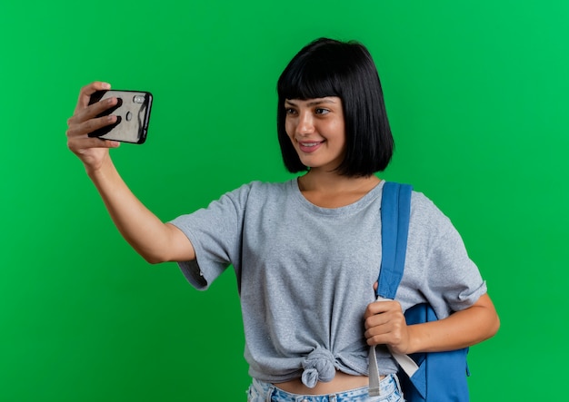 배낭을 입고 웃는 젊은 갈색 머리 백인 여자는 복사 공간이 녹색 배경에 고립 된 selfie를 복용 전화에서 보이는
