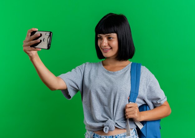 Smiling young brunette caucasian girl wearing backpack looks at phone taking selfie isolated on green background with copy space