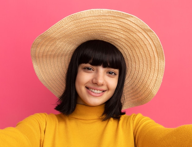 Smiling young brunette caucasian girl wearing beach hat pretends to hold camera taking selfie isolated on pink wall with copy space