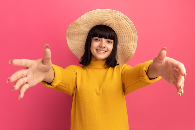 Smiling young brunette caucasian girl wearing beach hat stretching out hands isolated on pink wall with copy space