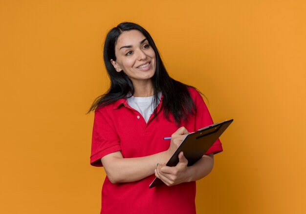 Smiling young brunette caucasian girl wearing red shirt holds pen and clipboard looking at side isolated on orange wall