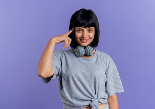 Smiling young brunette caucasian girl with headphones around neck puts finger on temple isolated on purple background with copy space