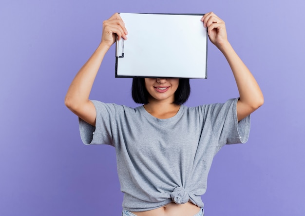 Smiling young brunette caucasian woman closes eyes holding clipboard isolated on purple background with copy space