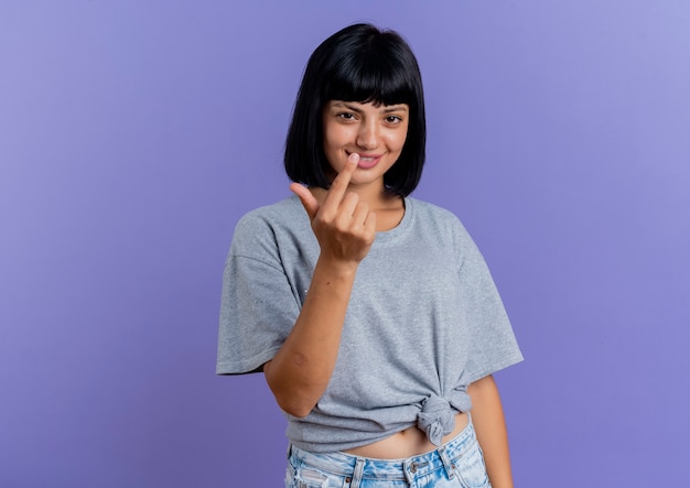 Smiling young brunette caucasian woman gestures come here with finger isolated on purple background with copy space