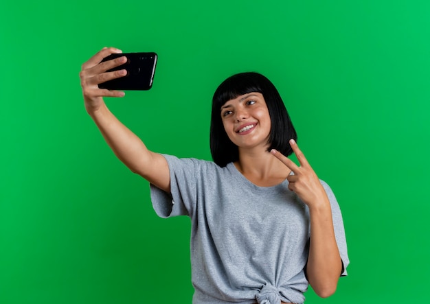 Smiling young brunette caucasian woman gestures victory hand sign looking at phone