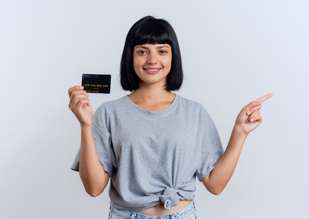 Улыбающаяся молодая брюнетка кавказская женщина держит кредитную карту и указывает в сторону