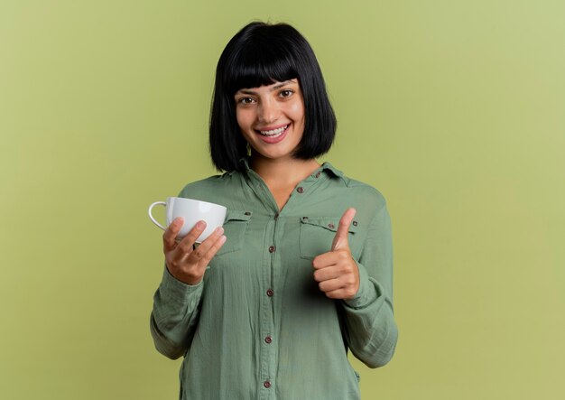 Smiling young brunette caucasian woman holds cup and thumbs up isolated on olive green background with copy space