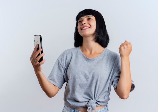 Smiling young brunette caucasian woman keeps fist and holds phone looking up Free Photo