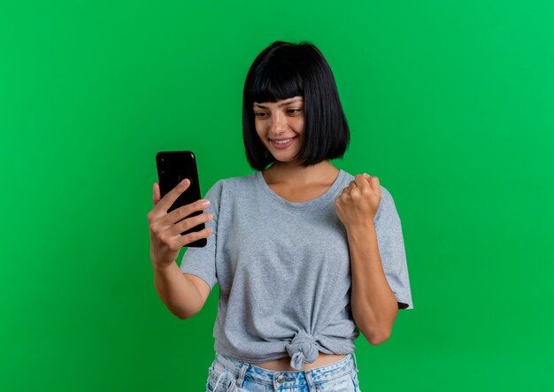 Smiling young brunette caucasian woman keeps fist and looks at phone