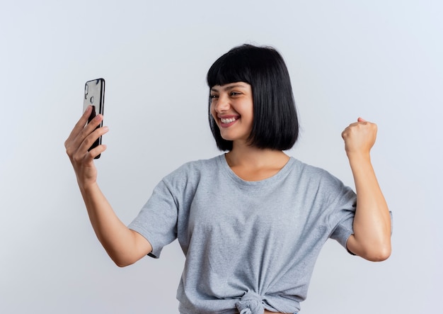 Smiling young brunette caucasian woman keeps fist and looks at phone