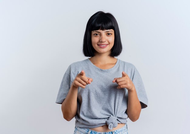 Smiling young brunette caucasian woman points at camera with two handsisolated