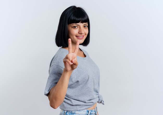 Smiling young brunette caucasian woman stands sideways gesturing victory hand sign