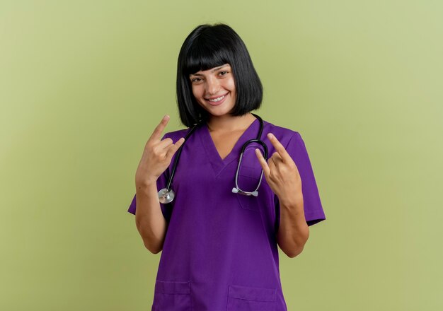 Smiling young brunette female doctor in uniform with stethoscope gestures horns hand sign with two hands 