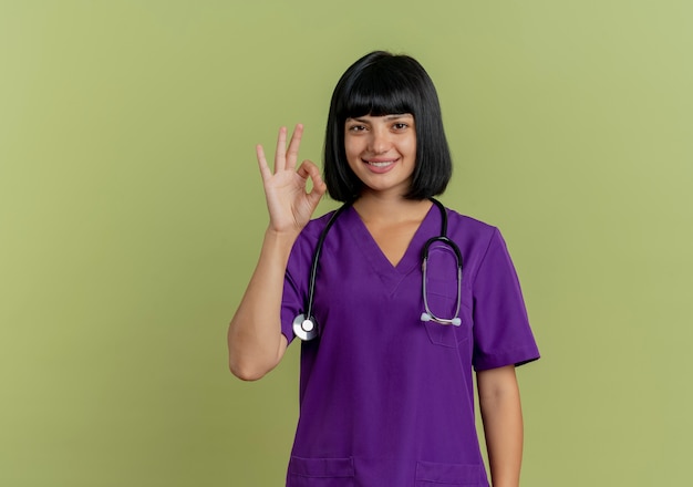 Smiling young brunette female doctor in uniform with stethoscope gestures ok hand sign isolated on olive green background with copy space