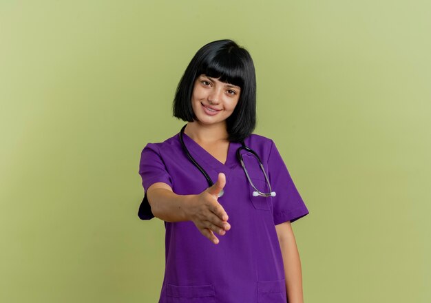 Smiling young brunette female doctor in uniform with stethoscope holds out hand 