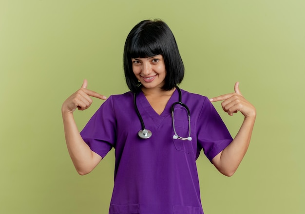 Sorridente giovane dottoressa bruna in uniforme con lo stetoscopio punta a se stessa con due mani isolate su sfondo verde oliva con spazio di copia