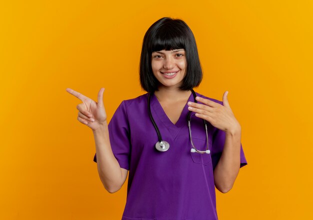 Smiling young brunette female doctor in uniform with stethoscope points at side isolated on orange background with copy space
