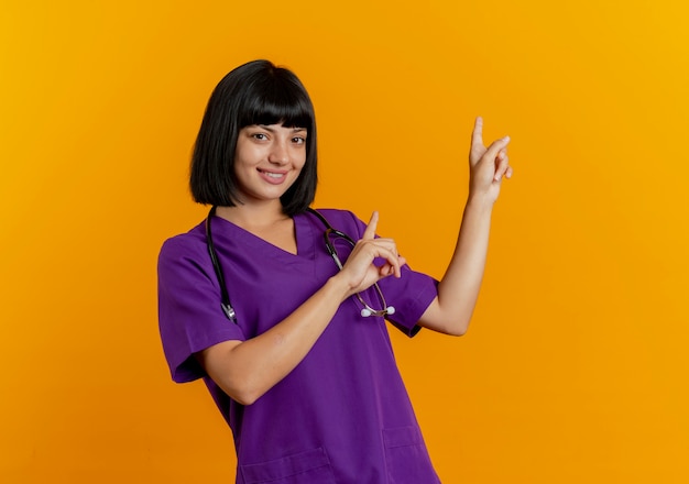 Foto gratuita sorridente giovane donna bruna medico in uniforme con i punti dello stetoscopio dietro con due mani