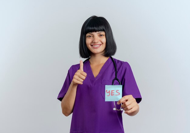 Улыбающаяся молодая брюнетка женщина-врач в униформе со стетоскопом показывает палец вверх и держит примечание да