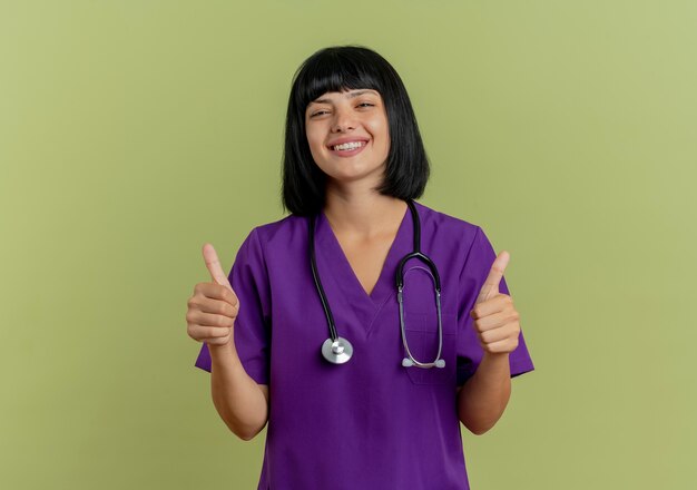 Улыбающаяся молодая брюнетка женщина-врач в униформе со стетоскопом показывает палец вверх двумя руками, изолированными на оливково-зеленом фоне с копией пространства