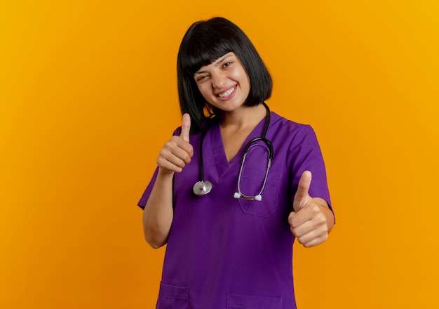 Улыбающаяся молодая брюнетка женщина-врач в униформе со стетоскопом показывает палец вверх двумя руками, изолированными на оранжевом фоне с копией пространства