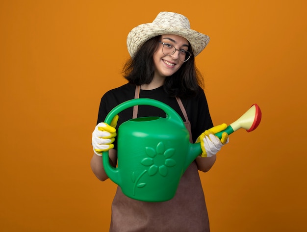 Бесплатное фото Улыбающаяся молодая брюнетка женщина-садовник в оптических очках и в униформе в садовой шляпе и перчатках держит лейку, изолированную на оранжевой стене