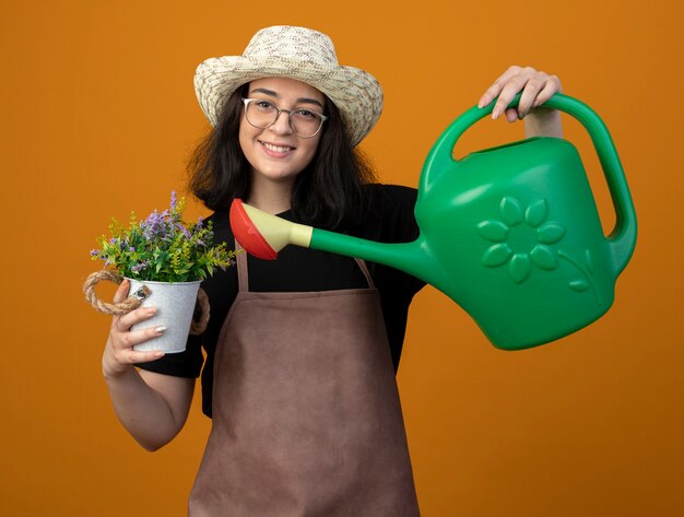 Smiling young brunette female gardener in optical glasses and in uniform wearing gardening hat holding watering can over flowerpot isolated on orange wall with copy space