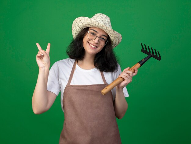 Il giovane giardiniere femminile castana sorridente in vetri ottici e cappello da giardinaggio d'uso uniforme tiene il segno della mano di vittoria di rastrello e gesti isolato sulla parete verde