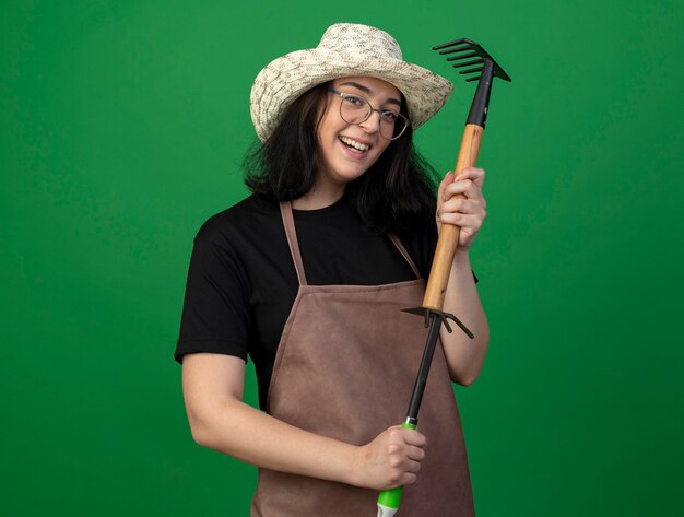 Smiling young brunette female gardener in optical glasses and uniform wearing gardening hat holds rake over hoe rake isolated on green wall