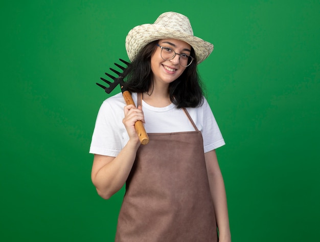 Il giovane giardiniere femminile castana sorridente in vetri ottici e cappello da giardinaggio d'uso uniforme tiene il rastrello sulla spalla isolata sulla parete verde